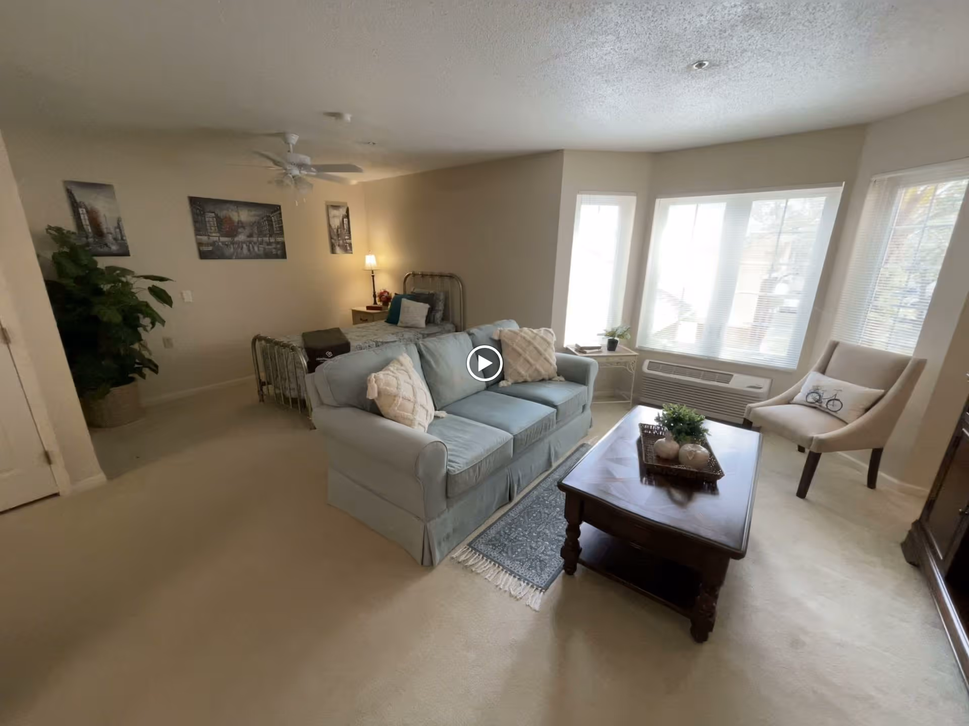 Studio-style living room with a sofa and coffee table in front of large bay windows and a bed visible in the background.