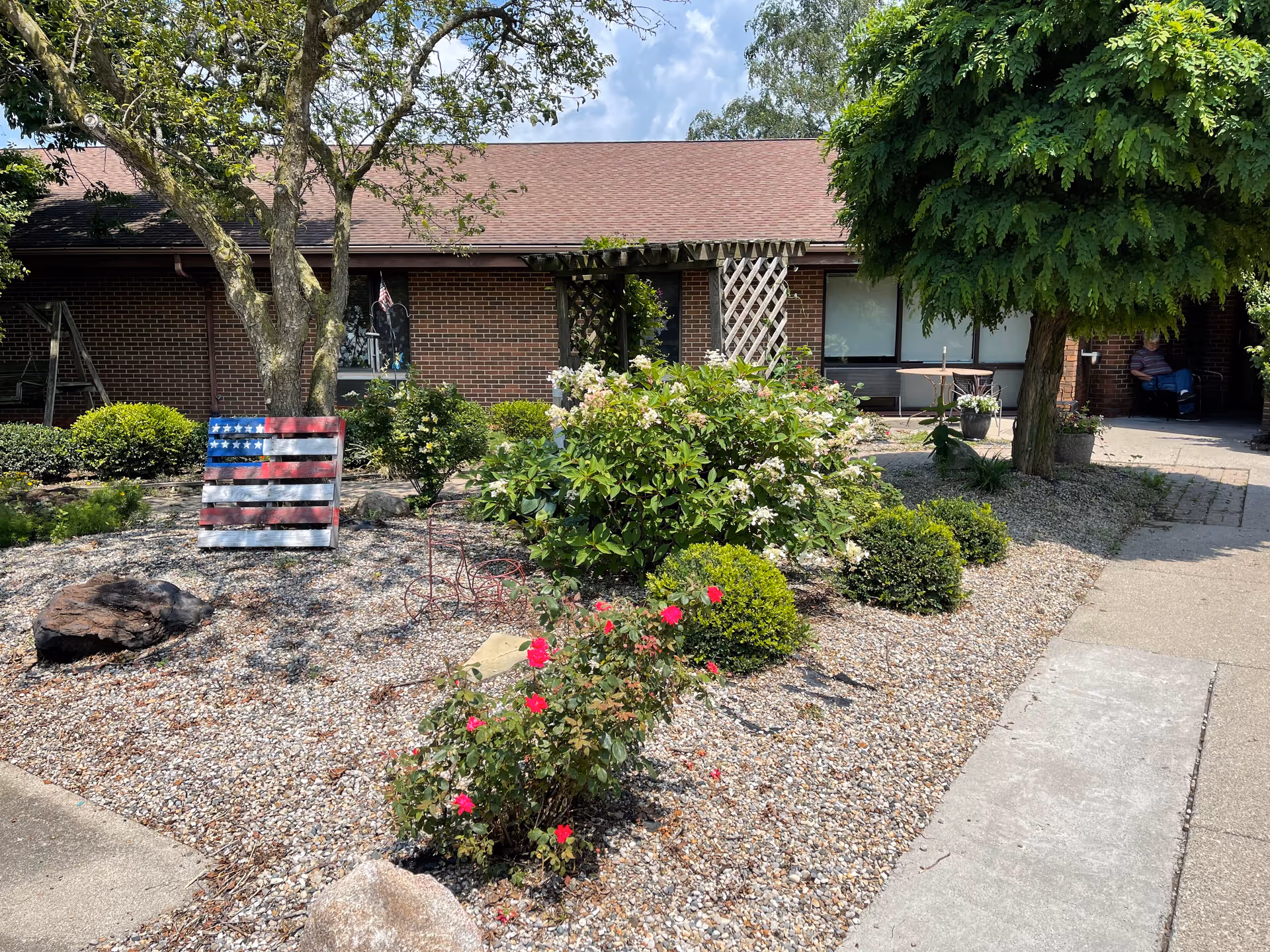Outdoor garden area in front of a brick building with a brown roof. The garden features various bushes, flowering plants, a tree, and a decorative American flag made from wooden slats. There is a paved walkway on the right side and a person sitting in the shaded area near the building entrance.