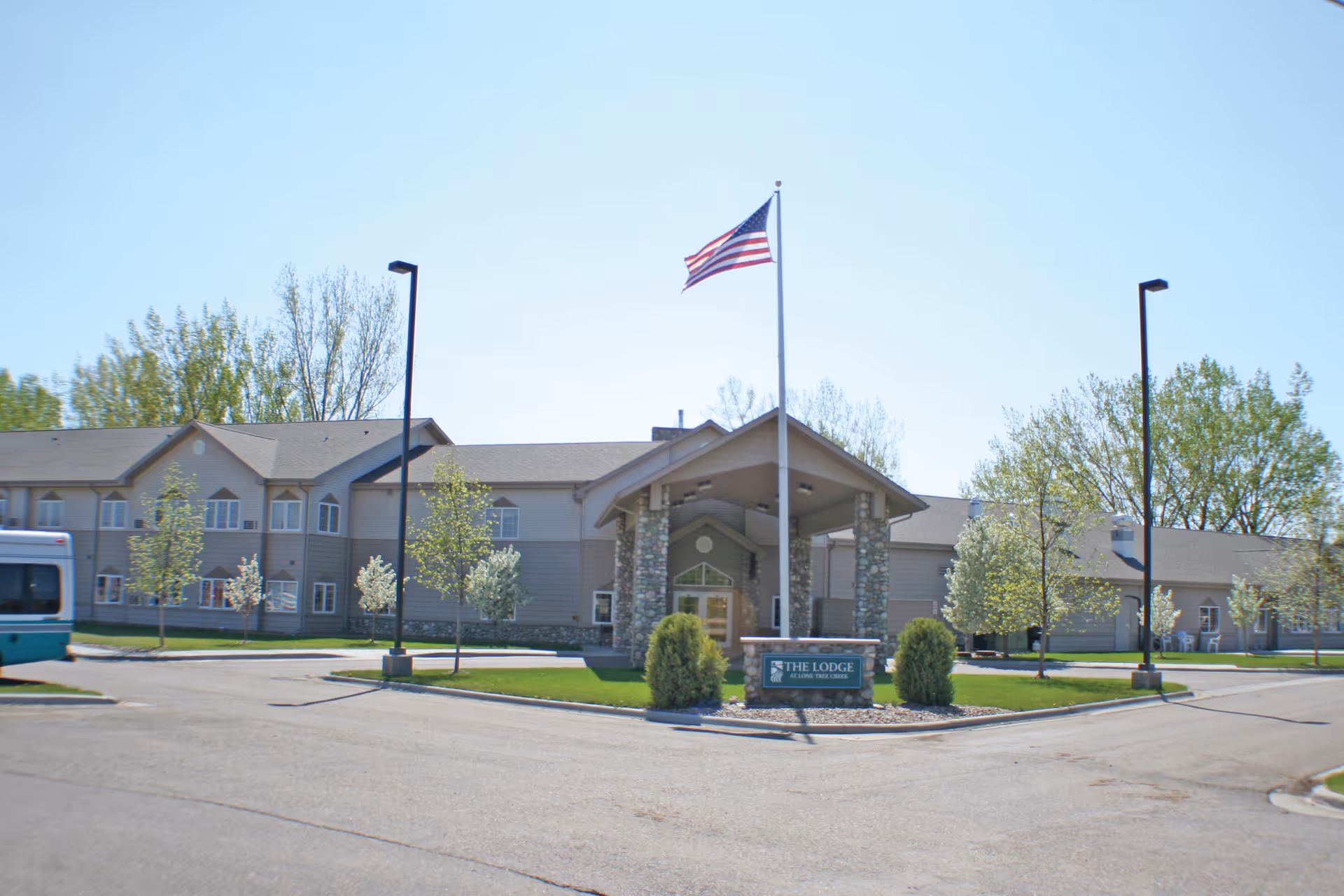 Exterior view of The Lodge - Managed by Sidney Health Center, a two-story building with a covered entrance supported by stone pillars, an American flag flying on a flagpole in front, surrounded by small trees and a paved driveway.