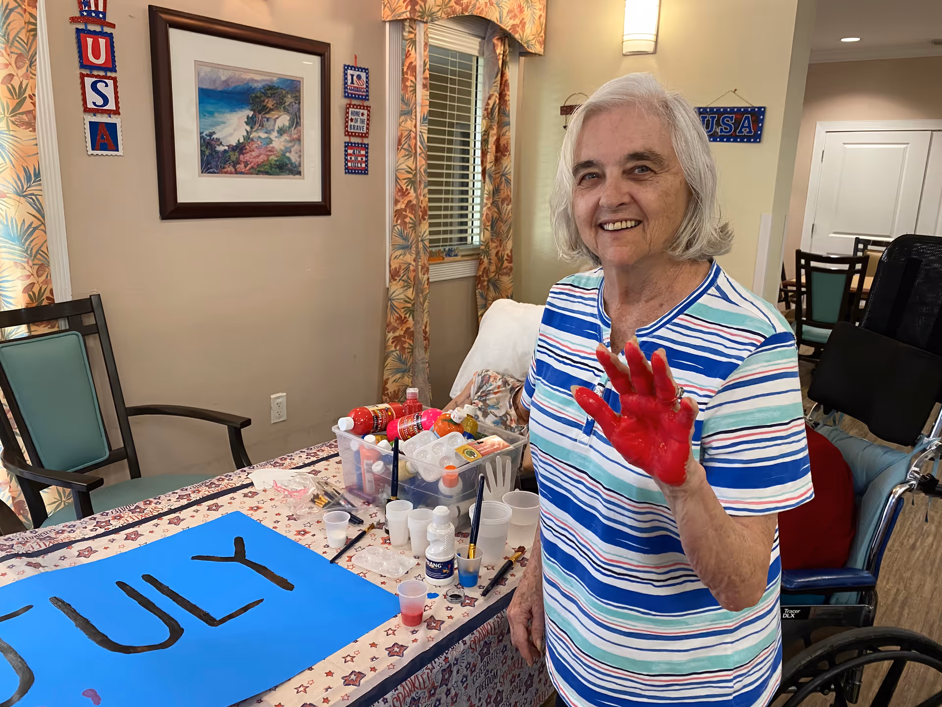 An elderly woman with gray hair wearing a striped shirt is smiling and showing her right hand covered in red paint. She is standing next to a table with art supplies, including paint bottles, brushes, and cups. A blue poster with the word 'JULY' partially visible is on the table. The room has patriotic decorations with USA-themed banners and a framed painting on the wall.