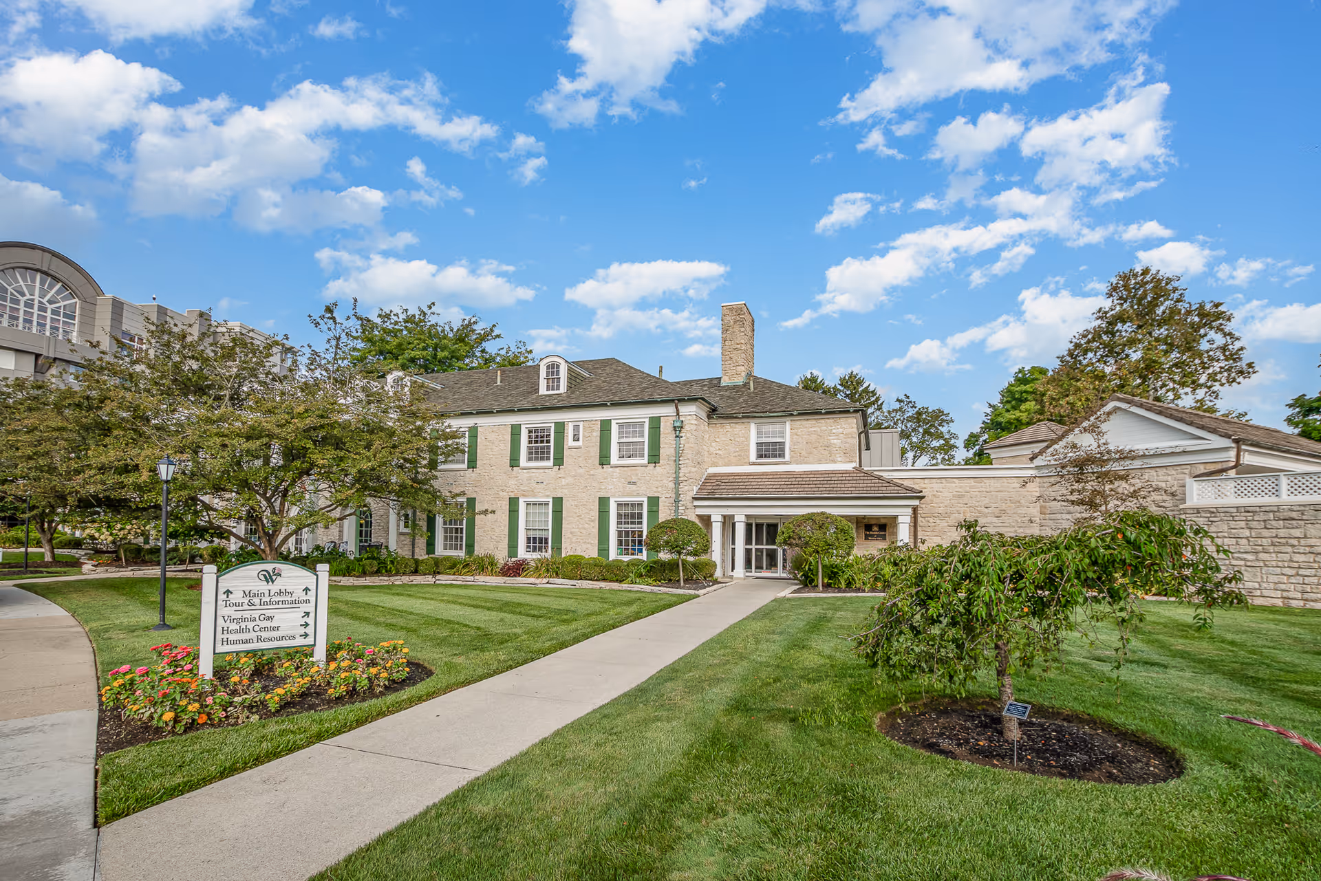 Front exterior of a senior living building with a paved walkway, manicured lawn, landscaping, and a clear blue sky.