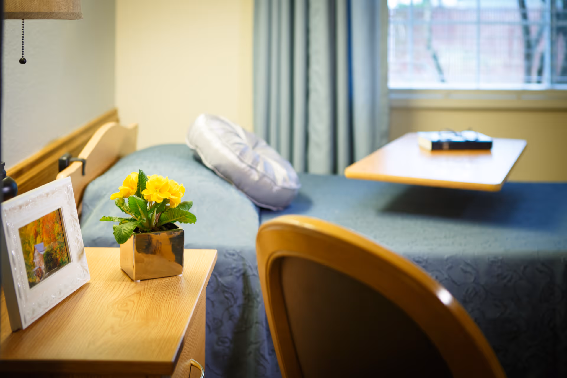 A tidy bedroom with a single bed, bedside table holding a potted yellow flower and framed photo, a chair, and a window.