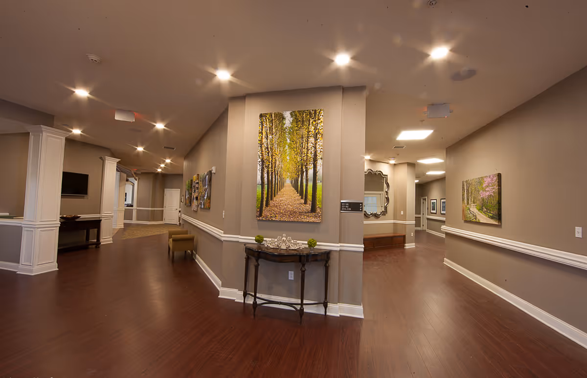 Interior hallway of a senior living facility with wooden flooring, beige walls, and white trim. The hallway features several pieces of wall art including a large photo of a tree-lined path, a decorative mirror, and a small table with decorative items. The ceiling has recessed lighting and there are multiple doorways leading to other rooms.