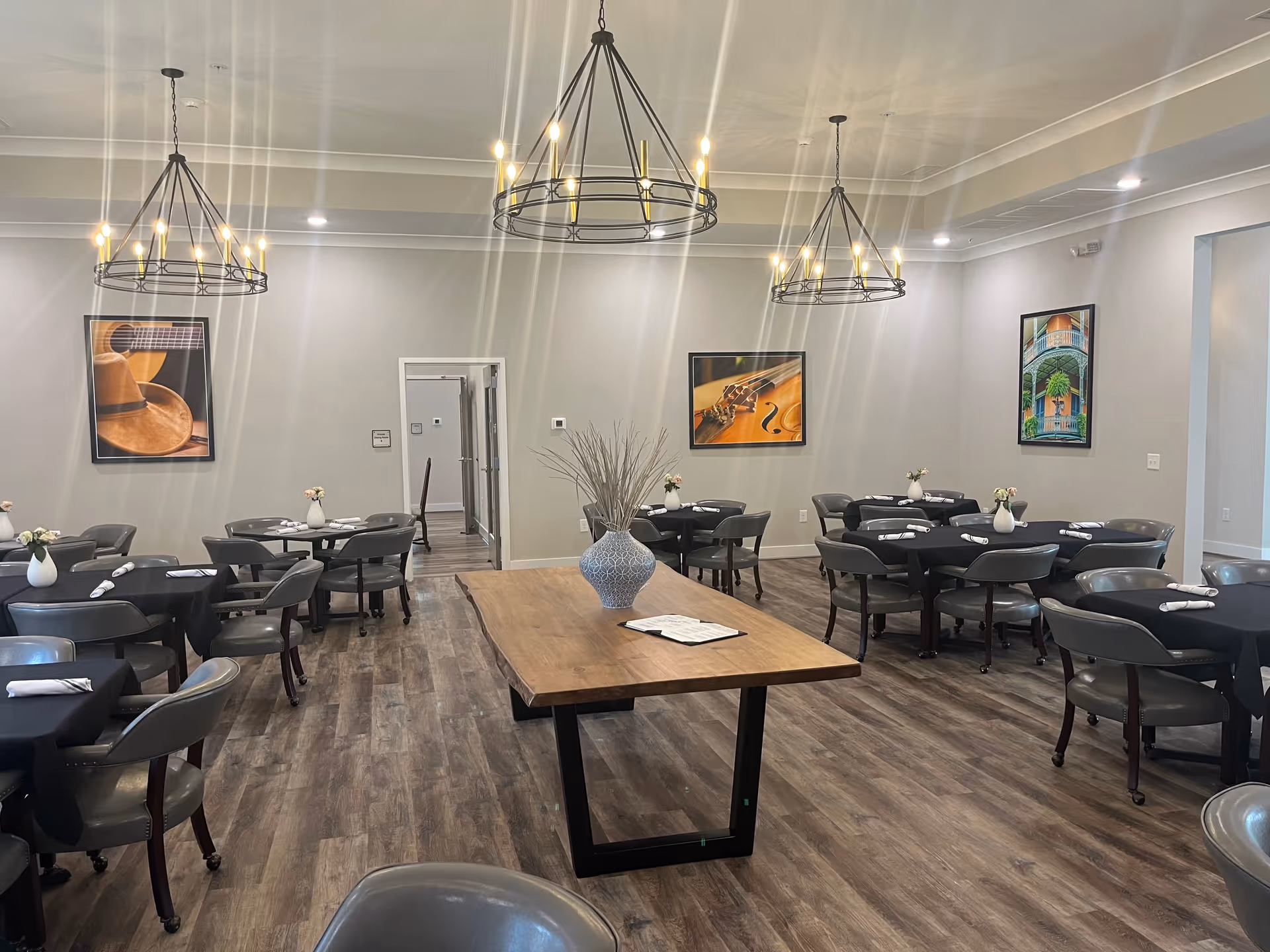A dining room with multiple round tables and black chairs arranged neatly. Each table has a small white vase with flowers and rolled white napkins. Three modern black chandeliers with candle-style lights hang from the ceiling. The floor is wood, and the walls are light gray with three framed pictures featuring musical instruments and a colorful building. A rectangular wooden table with a blue vase and dried plants is centered in the room.