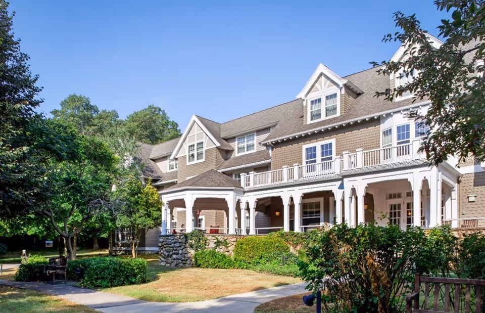 Exterior view of a large, multi-story senior living facility building with beige siding and white trim. The building features a covered porch with columns and a balcony above. Surrounding the building are green trees, bushes, and a well-maintained lawn under a clear blue sky.