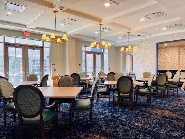 A spacious dining room with multiple wooden tables and green cushioned chairs arranged neatly. The room features large windows and glass doors allowing natural light to enter. Several chandeliers with yellow lampshades hang from the ceiling, and the carpet has a dark blue patterned design.