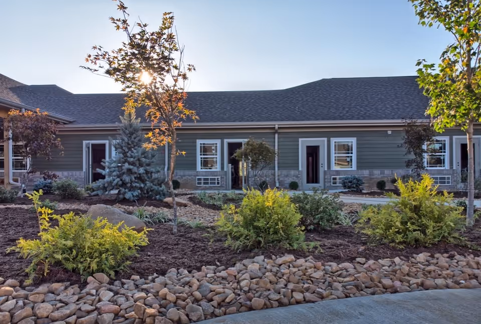 Exterior view of a single-story building with green siding and white-trimmed windows and doors. The foreground features a landscaped garden with small trees, shrubs, and rocks under a clear blue sky with the sun shining through the branches of a tree.