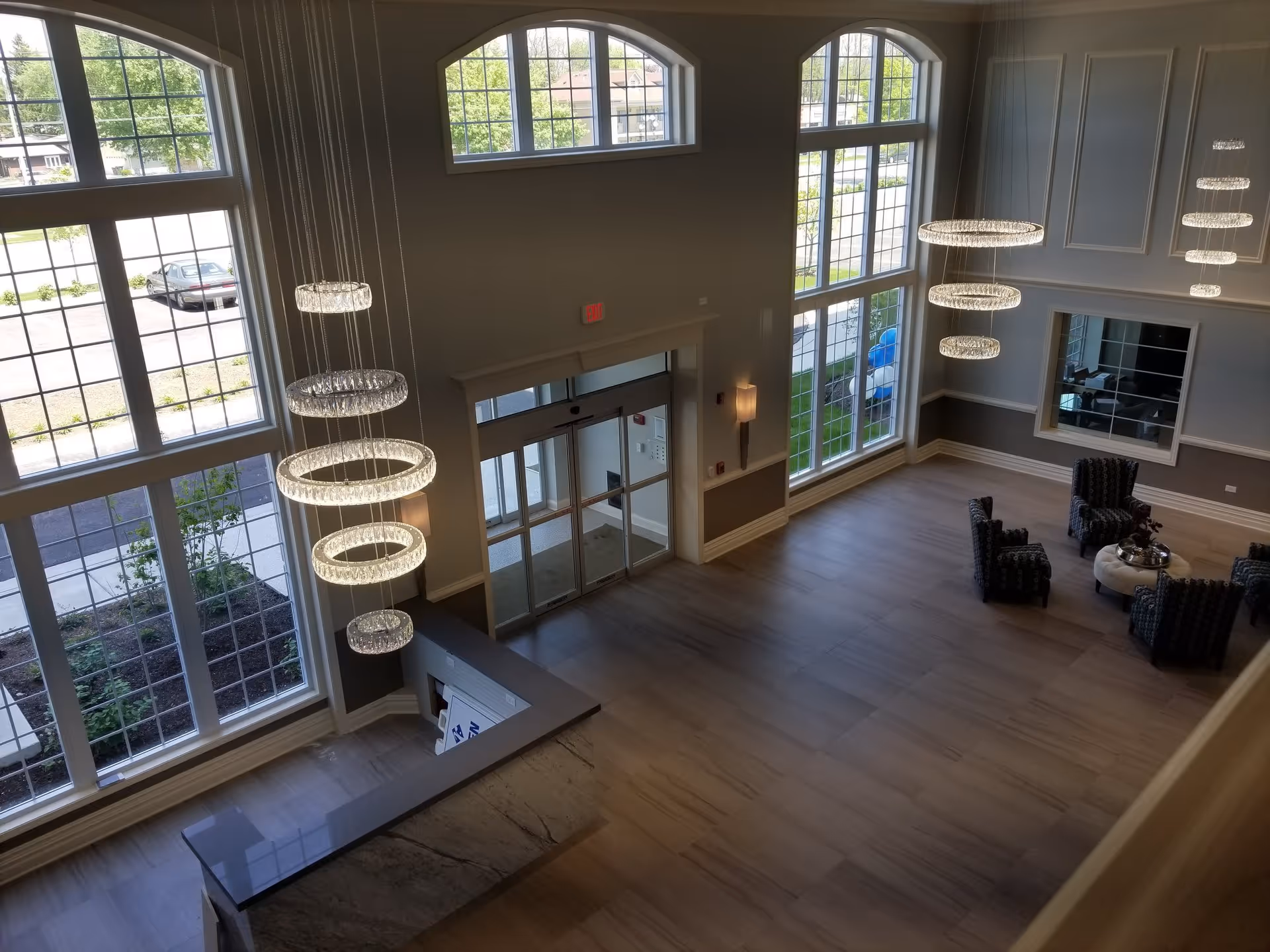 Spacious and well-lit lobby area of The Radcliff facility with large floor-to-ceiling windows, modern circular hanging light fixtures, a reception desk, and a seating area with four patterned armchairs around a round table.