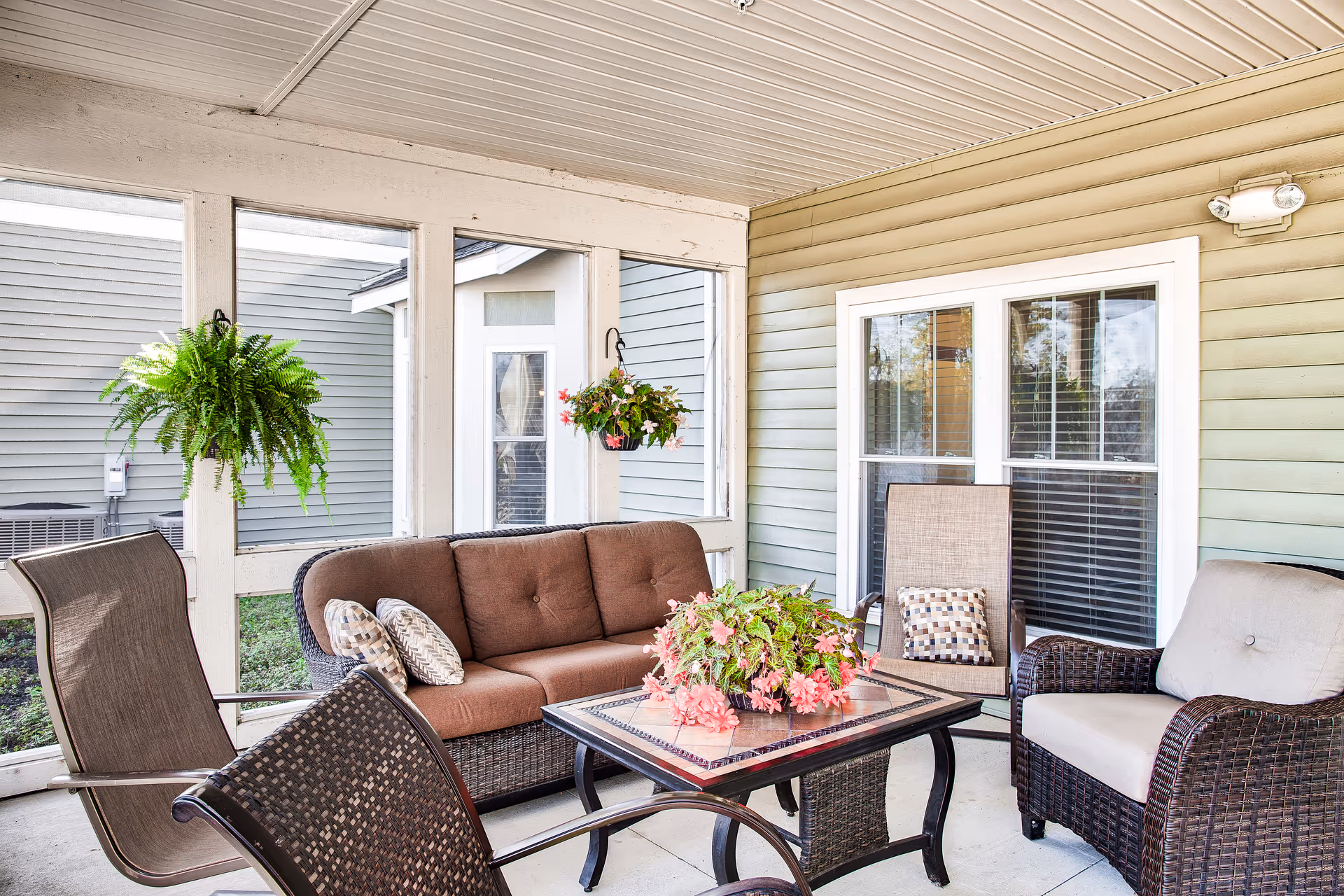 A covered outdoor patio area with wicker furniture including a brown cushioned sofa, two armchairs, and two additional chairs around a square table with a floral centerpiece. Hanging plants are visible near the screened windows, and the exterior of the building with light green siding and white trim is seen in the background.