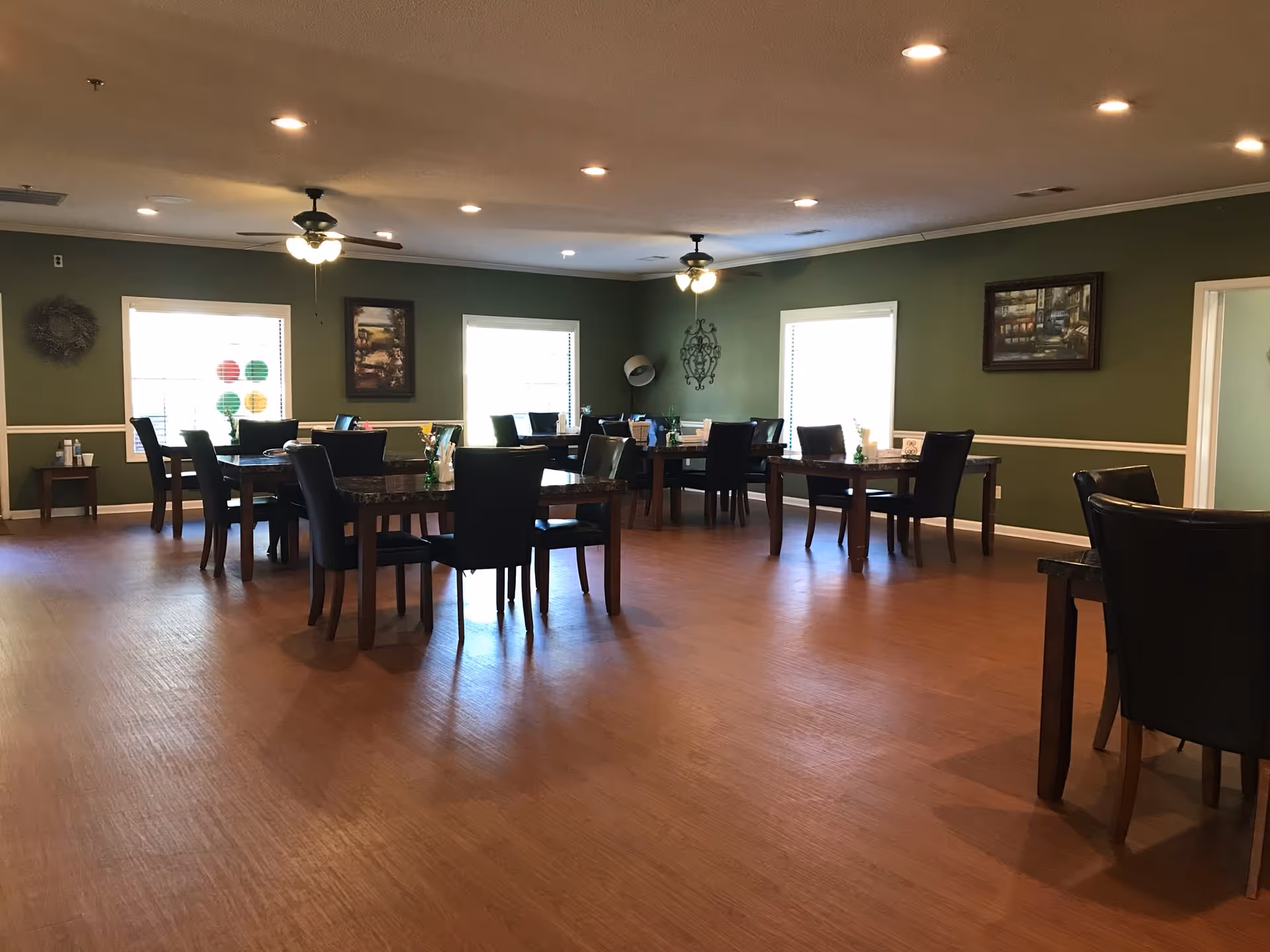 A dining room with several dark wooden tables and black chairs arranged neatly on a wooden floor. The walls are painted green and decorated with framed paintings and wall art. Ceiling fans with lights are mounted on the ceiling, and windows with blinds allow natural light to enter the room.