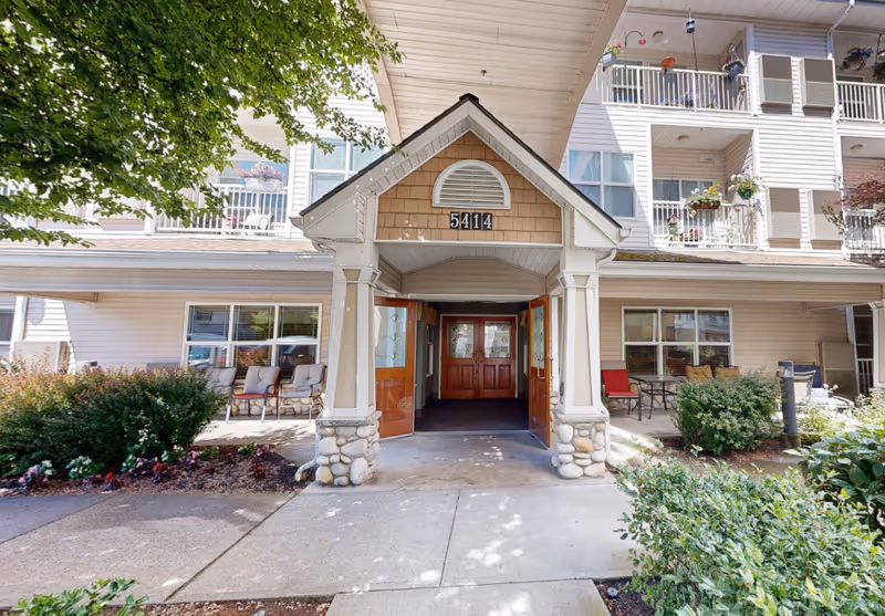 Entrance of a senior living facility with a covered porch supported by stone pillars, double wooden doors, and the number 5414 displayed above. The building has multiple balconies with potted plants and outdoor seating areas surrounded by greenery and flowers.