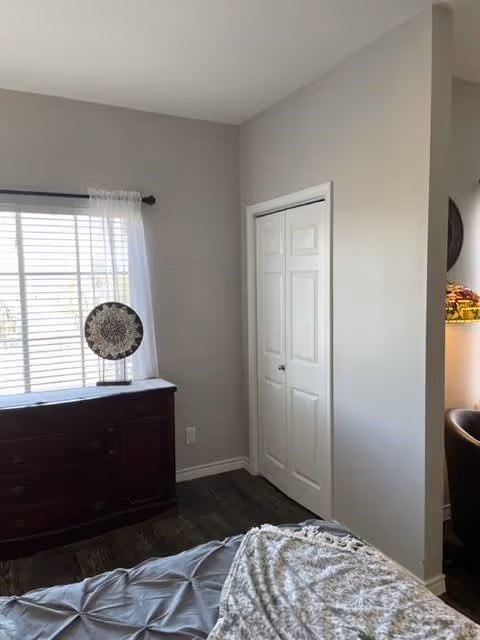A bedroom corner with a window covered by white sheer curtains, a dark wooden dresser with a decorative plate on top, a closed white closet door, and part of a bed with a gray comforter and patterned blanket.