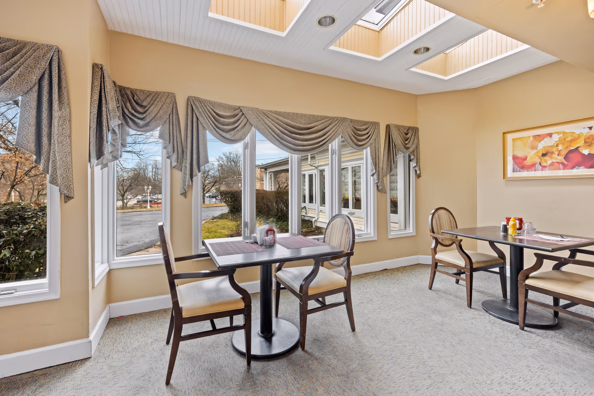 A bright dining area with two tables, each surrounded by two wooden chairs with beige cushions. The room has large windows with decorative draped curtains, allowing natural light to fill the space. The walls are painted a warm beige color, and there is a colorful floral painting hanging on one wall. The ceiling features skylights, enhancing the natural lighting in the room.