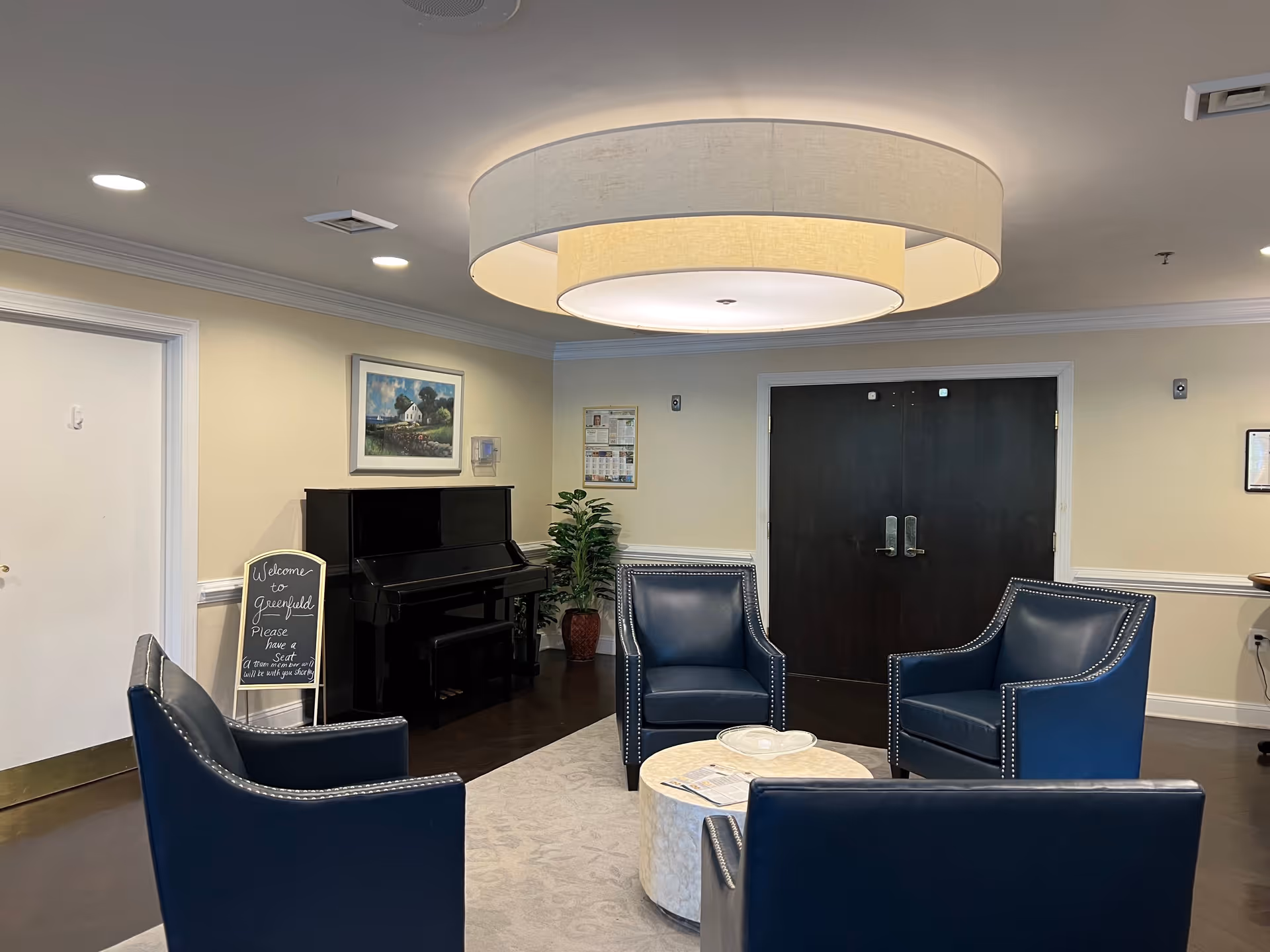 A cozy sitting area in a senior living facility with four blue armchairs arranged around a round white coffee table. A black upright piano is against the wall with a framed painting above it. A small chalkboard sign next to the piano reads 'Welcome to Greenfield Please have a seat.' The room has beige walls, dark double doors, a large circular ceiling light fixture, and a potted plant in the corner.