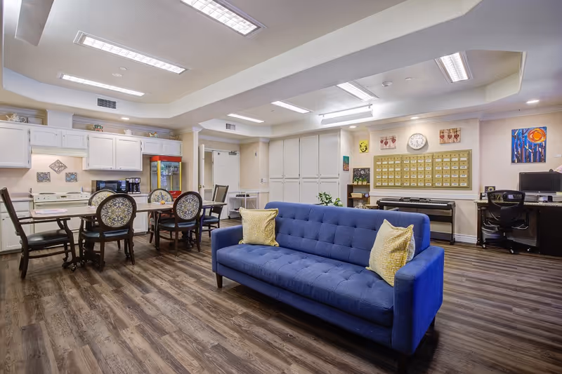 A spacious common area in a senior living facility featuring a blue couch with two yellow pillows in the foreground. Behind the couch, there is a dining table with six chairs, a kitchen area with white cabinets, a stove, and a coffee maker. On the right side, there is a desk with a computer and an office chair, a keyboard, and a wall-mounted mailbox organizer with a clock above it. The room has wood flooring and bright overhead lighting.
