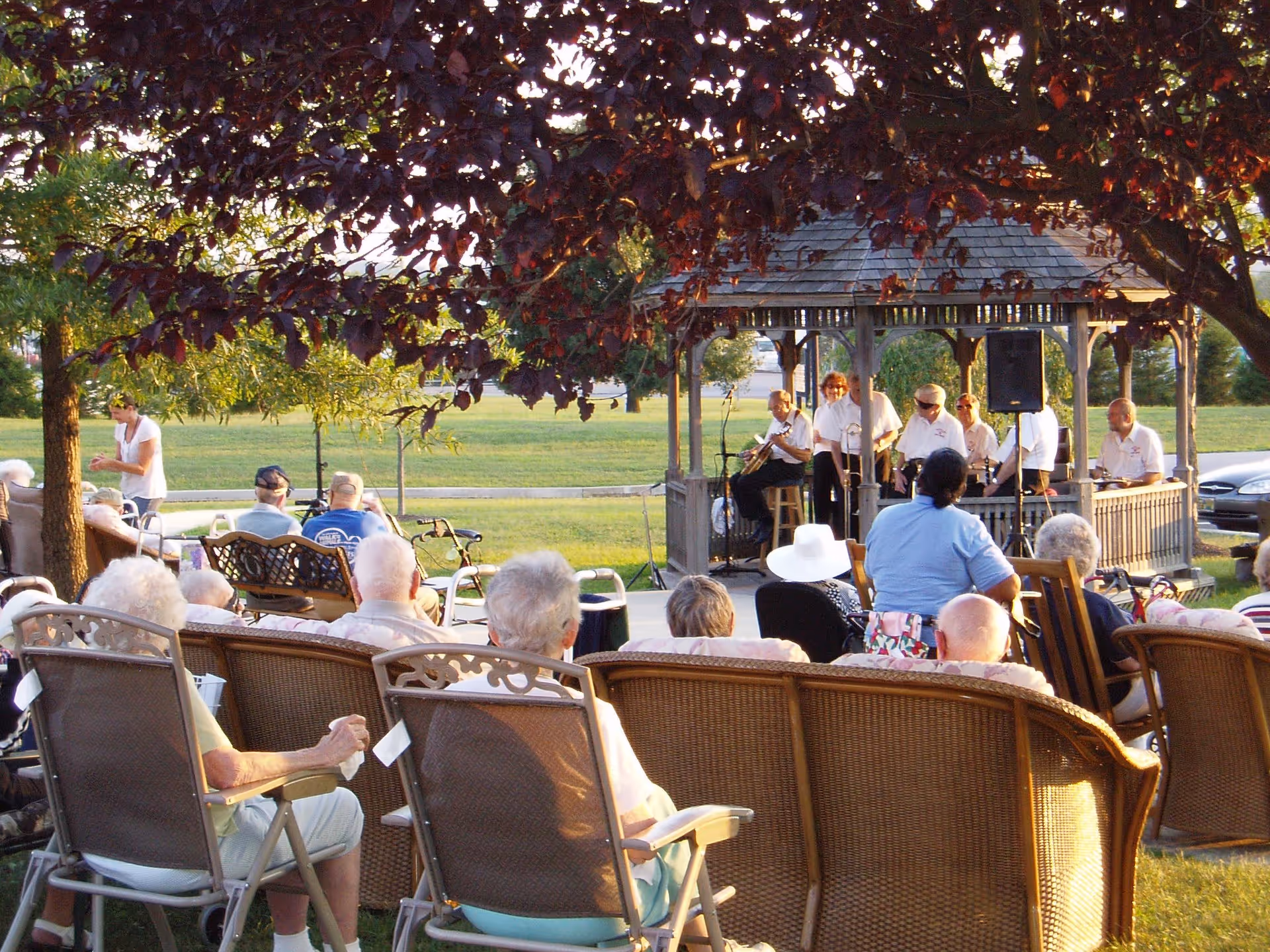 A group of elderly people seated outdoors on chairs and benches, watching a live band performing under a wooden gazebo. The scene is set in a grassy area with trees providing shade, and a few caregivers or staff members are present.