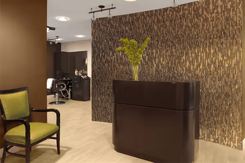 Reception area with a dark wood front desk, decorative mosaic wall, a green-upholstered chair, and salon stations visible in the background.