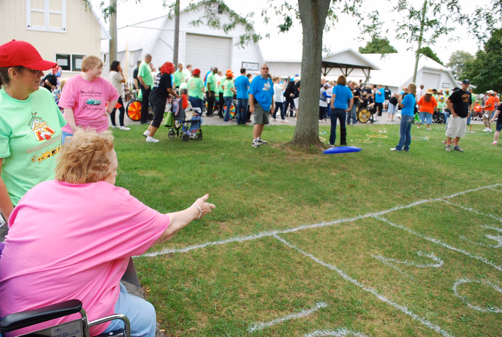 An outdoor event with many people gathered on a grassy area. In the foreground, an elderly woman in a pink shirt sitting in a wheelchair is reaching out to throw a ring or similar object. Other people, some wearing colorful shirts, are standing and watching. There are white chalk markings on the grass indicating game areas. Trees and white buildings are visible in the background.