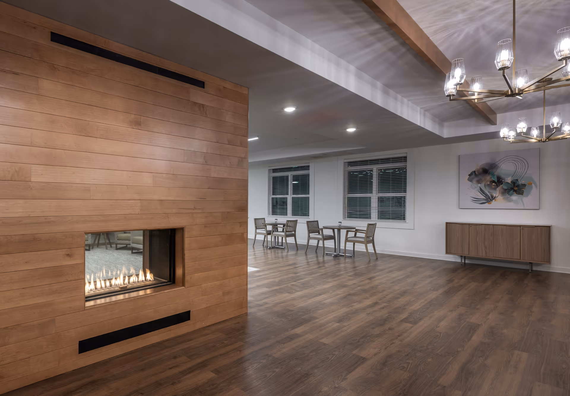 A modern interior space featuring a wooden paneled wall with a built-in gas fireplace. The room has wood flooring, a few small tables with chairs near windows with blinds, a wooden sideboard against the wall, and a contemporary chandelier with multiple lights hanging from the ceiling.