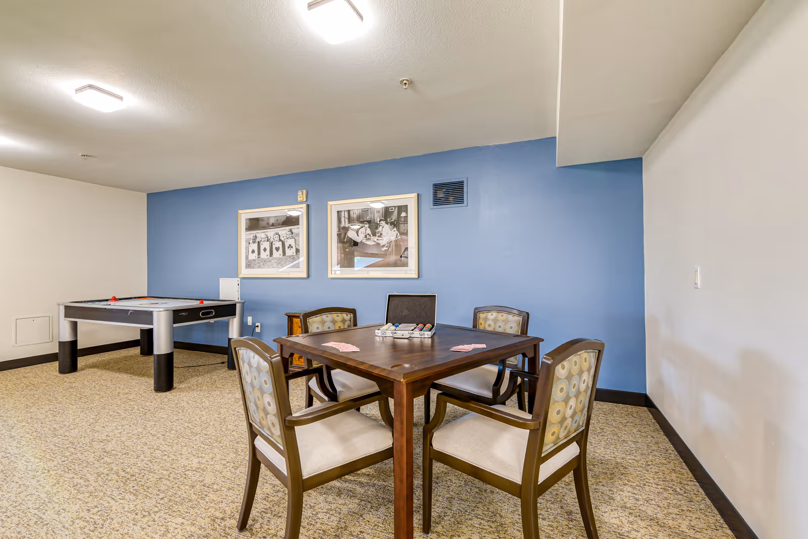 A recreation room with a square game table and four chairs in the foreground, an air hockey table to the left, and a blue accent wall with framed photos.