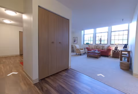 Interior view of a senior living facility showing a hallway with wood flooring leading to a living room area. The living room has a large window with multiple panes, a red sofa, two armchairs, a wooden coffee table, side tables with lamps, and a basket on the floor. The walls are light-colored, and there is a closet with wooden doors in the hallway.