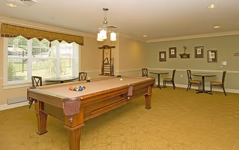A well-lit recreational room featuring a pool table with balls arranged for a game. The room has large windows with patterned valances, a wall-mounted cue rack, and several tables with chairs along the walls. The carpet is patterned and the walls are painted in neutral tones with framed artwork.