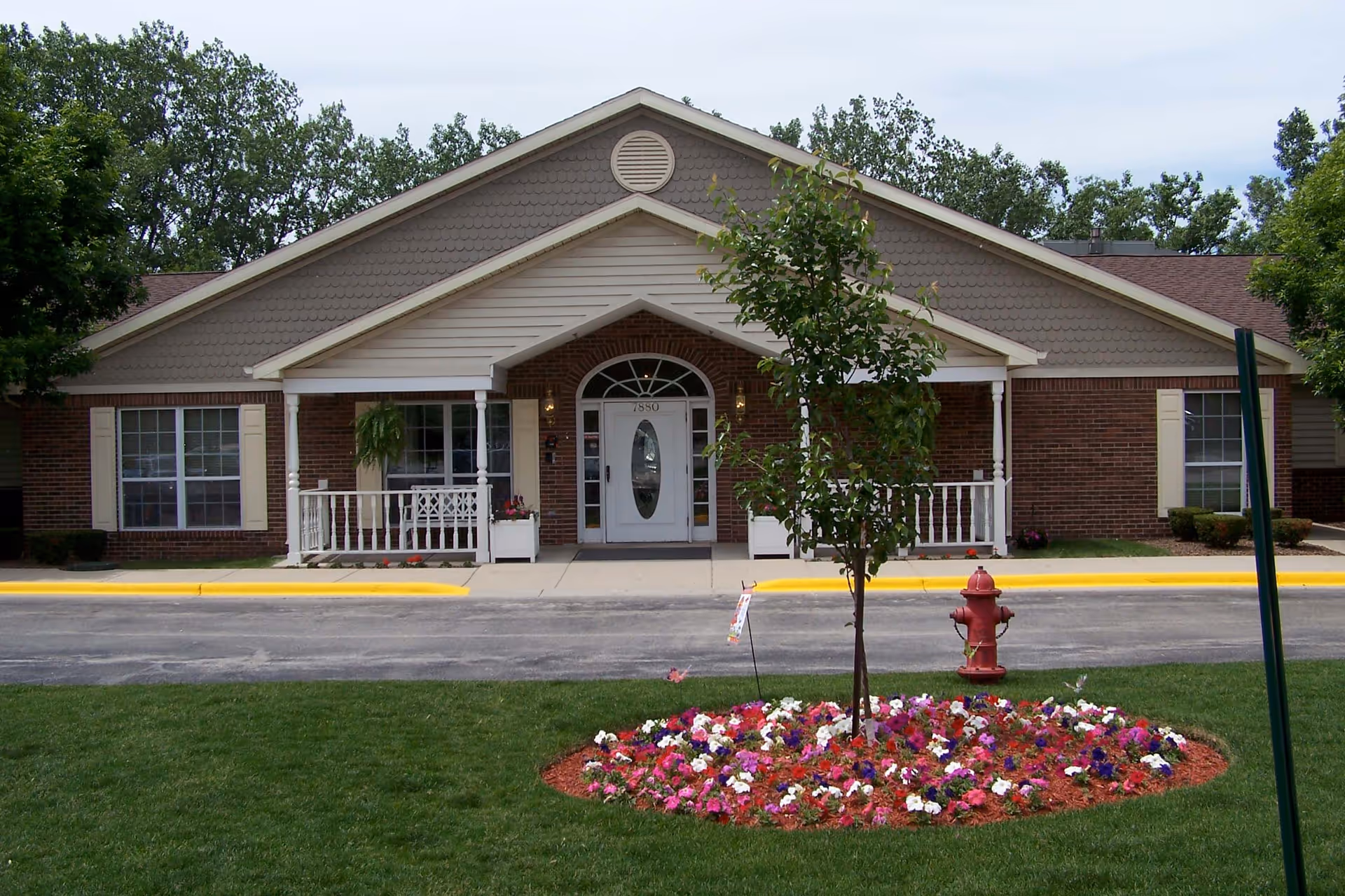 Front exterior view of a single-story brick building with a peaked roof, white trim, and a central entrance with a white door and sidelights. There is a small porch with white railings and potted plants on either side of the entrance. In front of the building is a circular flower bed with colorful flowers and a small tree, along with a red fire hydrant. The building is surrounded by green grass and trees in the background.
