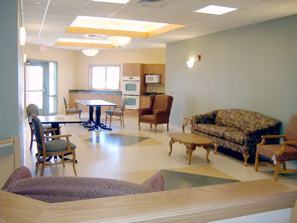 A bright and spacious common area with a kitchen in the background, featuring a stove, microwave, and cabinets. The room has several chairs and tables, a patterned sofa, armchairs, and a coffee table. The floor has a geometric pattern, and the ceiling has recessed lighting and hanging light fixtures.