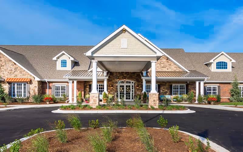 Front exterior view of Bickford of Shelby Township facility with a large covered entrance supported by white columns, stone and brick facade, multiple windows, and a landscaped circular driveway under a clear blue sky.