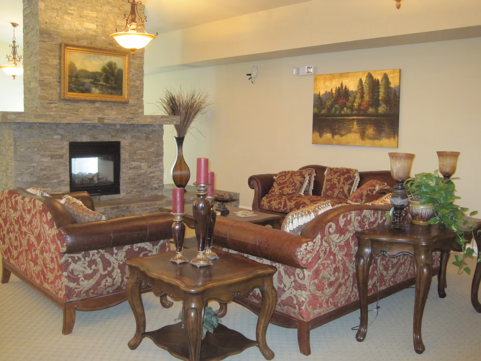 A cozy living room with two patterned red and brown sofas arranged around a stone fireplace. The room features wooden side tables with decorative candle holders and a potted plant. There are framed landscape paintings on the walls and warm lighting from ceiling fixtures.