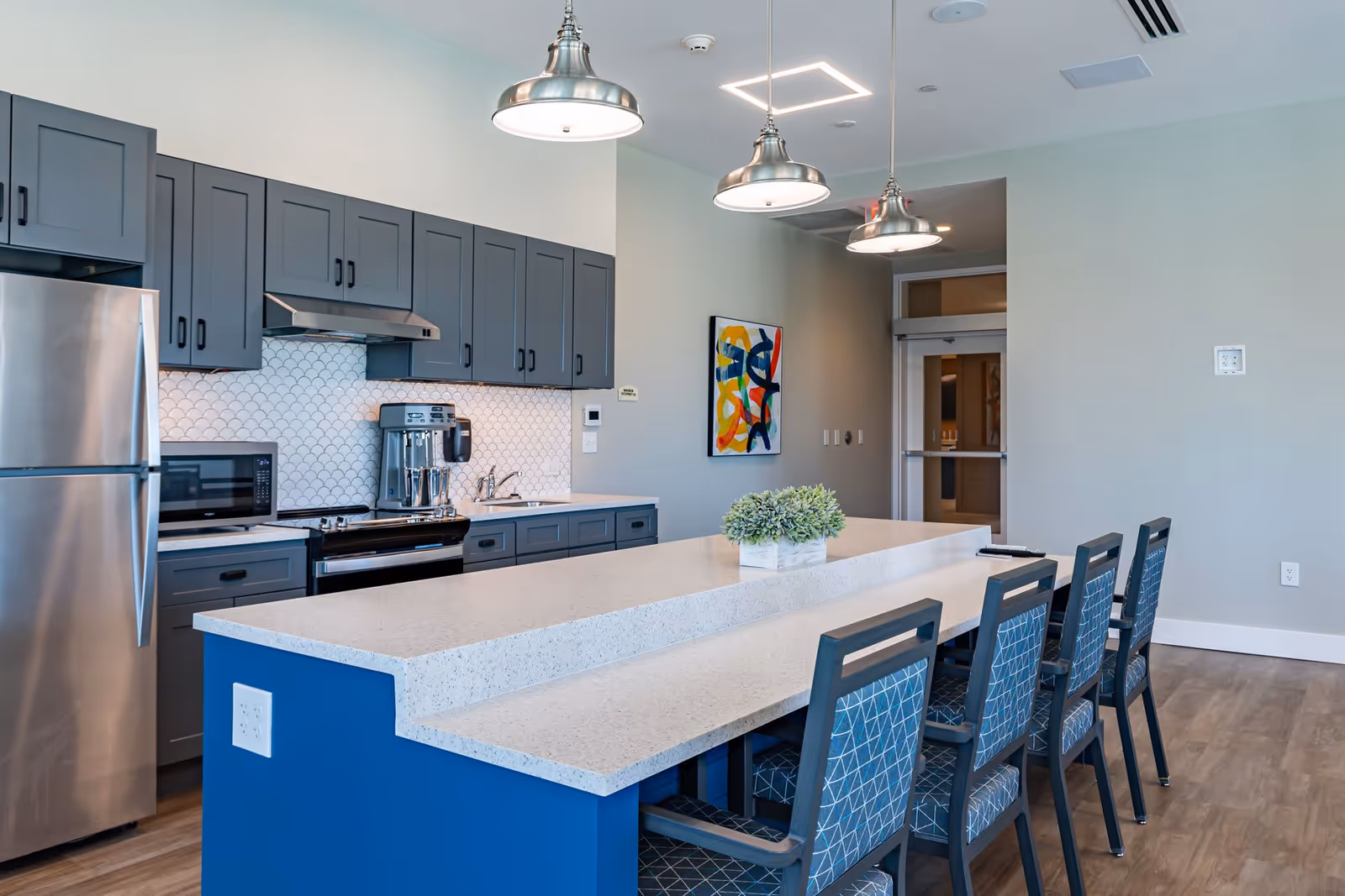 Modern kitchen area with blue lower cabinets and gray upper cabinets, a stainless steel refrigerator, microwave, stove, and coffee machine. A long white countertop island with four blue patterned chairs is in the foreground, with three hanging pendant lights above. A colorful abstract painting is on the wall near a doorway.