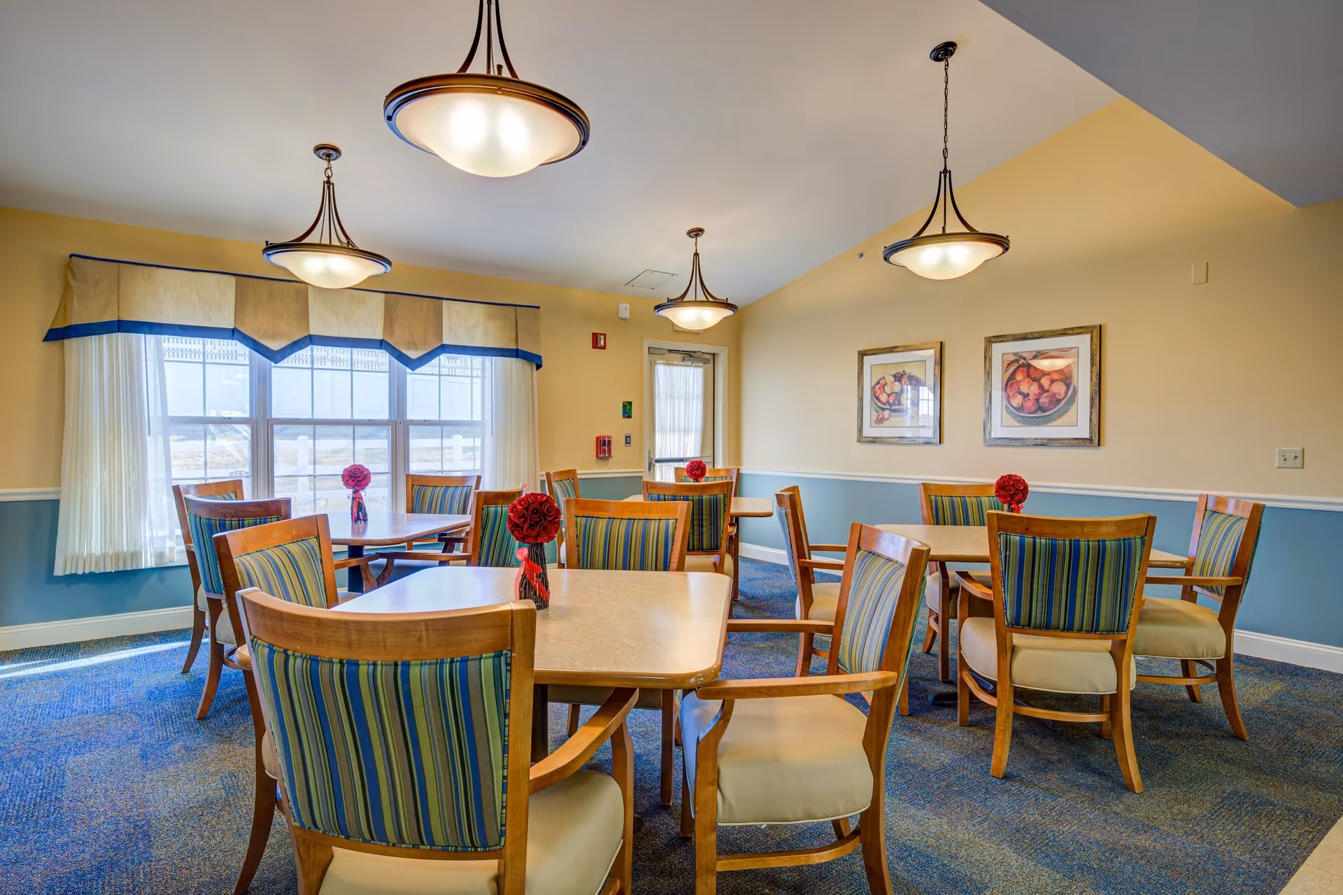 A bright dining room with several wooden tables and chairs featuring striped upholstery. Each table has a small red floral centerpiece. The room has large windows with sheer curtains and valances, blue and beige walls, carpeted floor, and three hanging ceiling lights. Two framed pictures of fruit bowls hang on one wall.