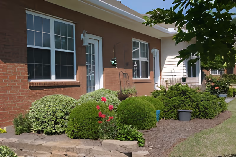 Front exterior of a single-story brick cottage with windows, a door, and manicured bushes and flowering shrubs.
