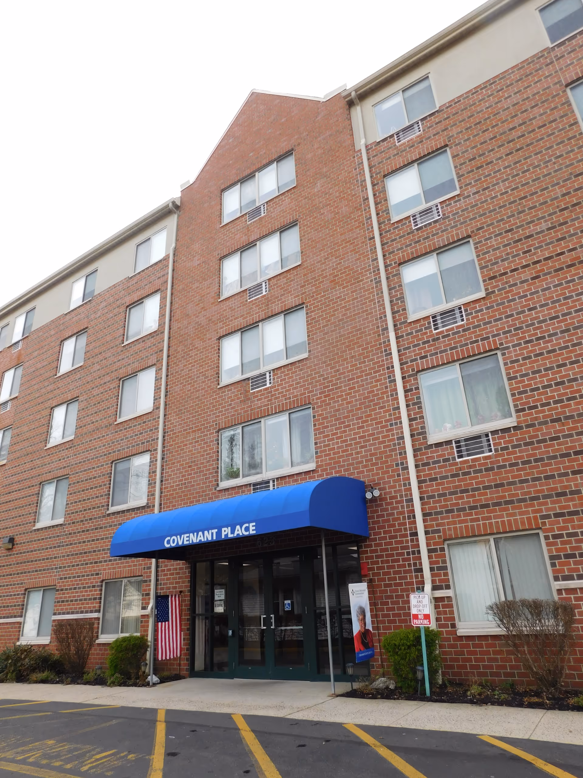 Exterior view of a multi-story brick building with a blue awning over the entrance that reads 'Covenant Place'. There are several windows on the building, a small American flag near the entrance, and some bushes along the front. A sign near the entrance indicates parking restrictions.