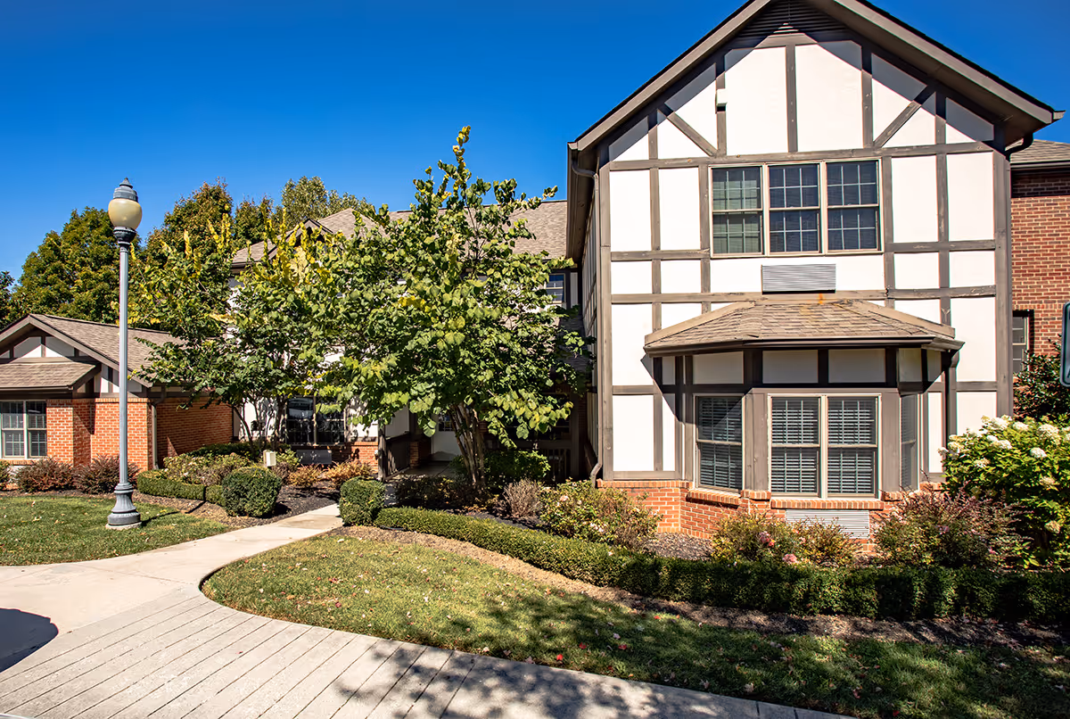 Exterior view of a senior living facility building with Tudor-style architecture, featuring white walls with dark wooden beams and a brick base. The building is surrounded by well-maintained landscaping including bushes, trees, and a green lawn. A sidewalk and a streetlamp are visible in the foreground under a clear blue sky.