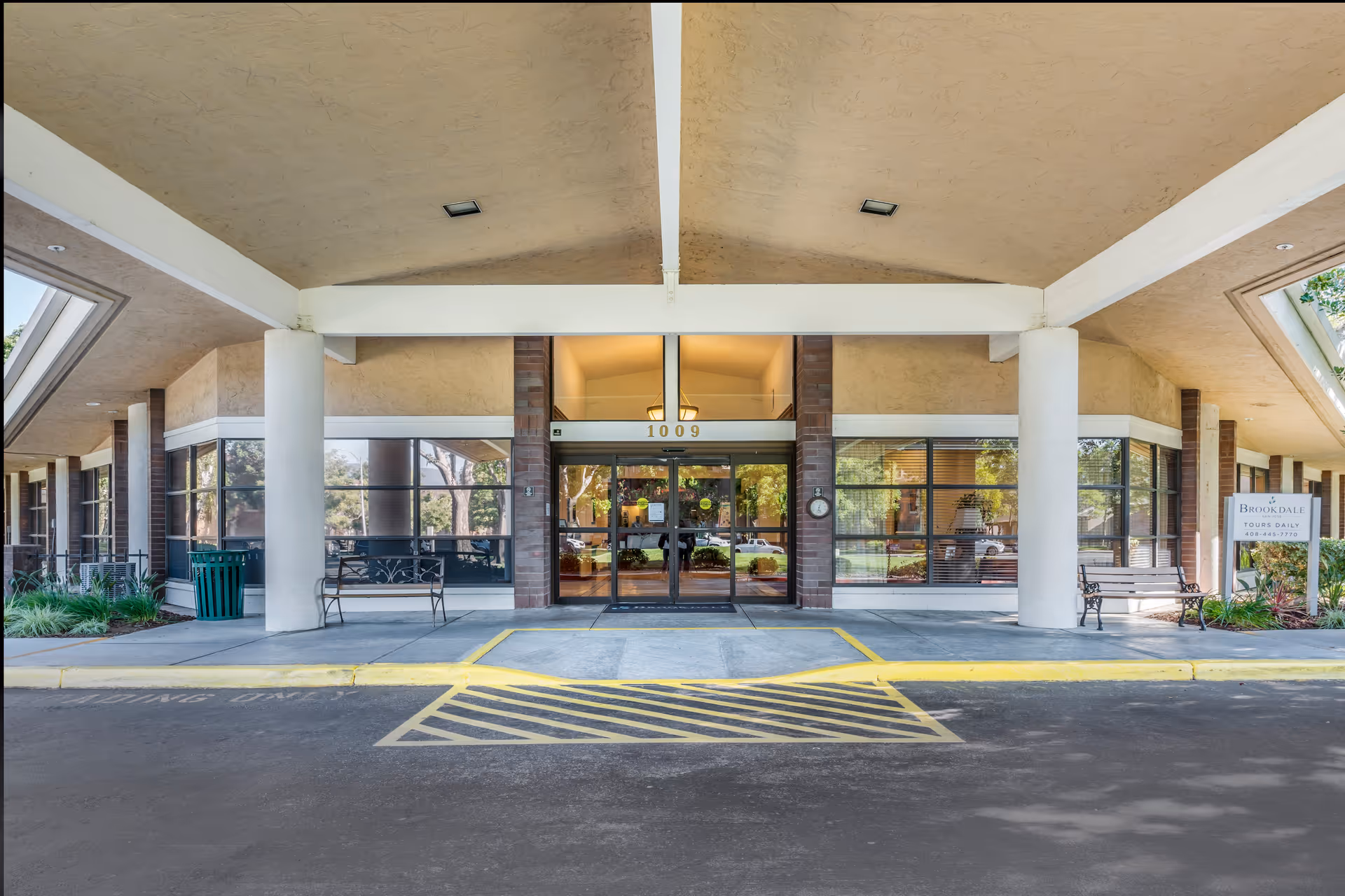 Front entrance of Brookdale San Jose facility with large glass doors, white pillars, benches on either side, and a covered driveway with yellow painted markings.