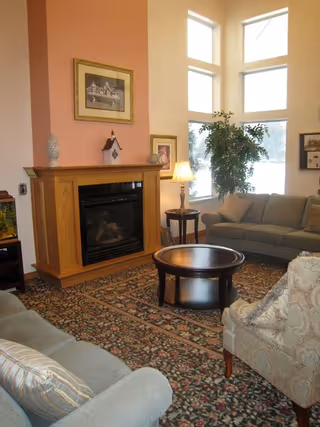 A cozy living room with a wooden fireplace, a round wooden coffee table, a patterned area rug, two sofas, an armchair, a side table with a lamp, framed artwork on the walls, and large windows letting in natural light.