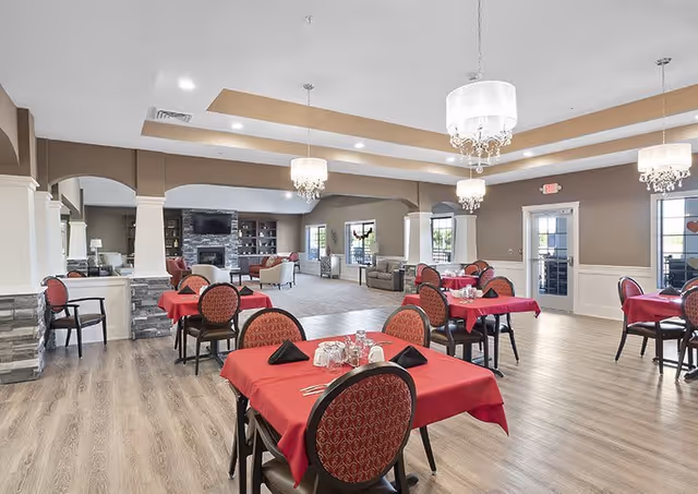 A spacious dining area with several tables covered in red tablecloths, each set with glasses, napkins, and silverware. The room features wooden flooring, elegant chandeliers hanging from a tray ceiling, and a seating area with sofas and a stone fireplace in the background.
