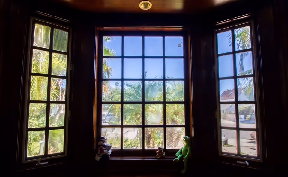 A bay window with three sections featuring wooden frames. Outside the window, palm trees and a clear blue sky are visible. On the window sill, there are three stuffed toys including a green frog, a small teddy bear, and a doll.