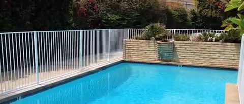 Outdoor swimming pool with clear blue water, surrounded by a white metal fence and a raised stone planter with greenery and plants in the background.