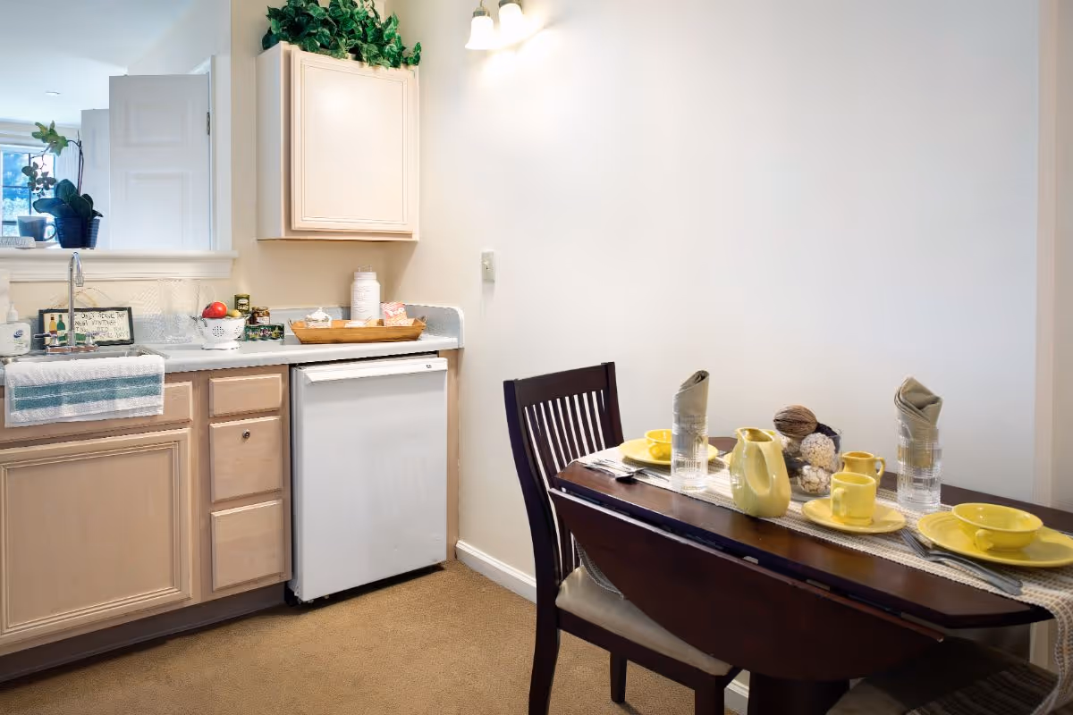 A small dining area and kitchen corner in a senior living facility. The dining table is set with yellow dishes, cups, and a pitcher, along with two glasses with folded napkins. The kitchen area has light wood cabinets, a white dishwasher, a sink with a towel hanging, and some decorative plants and items on the countertop.