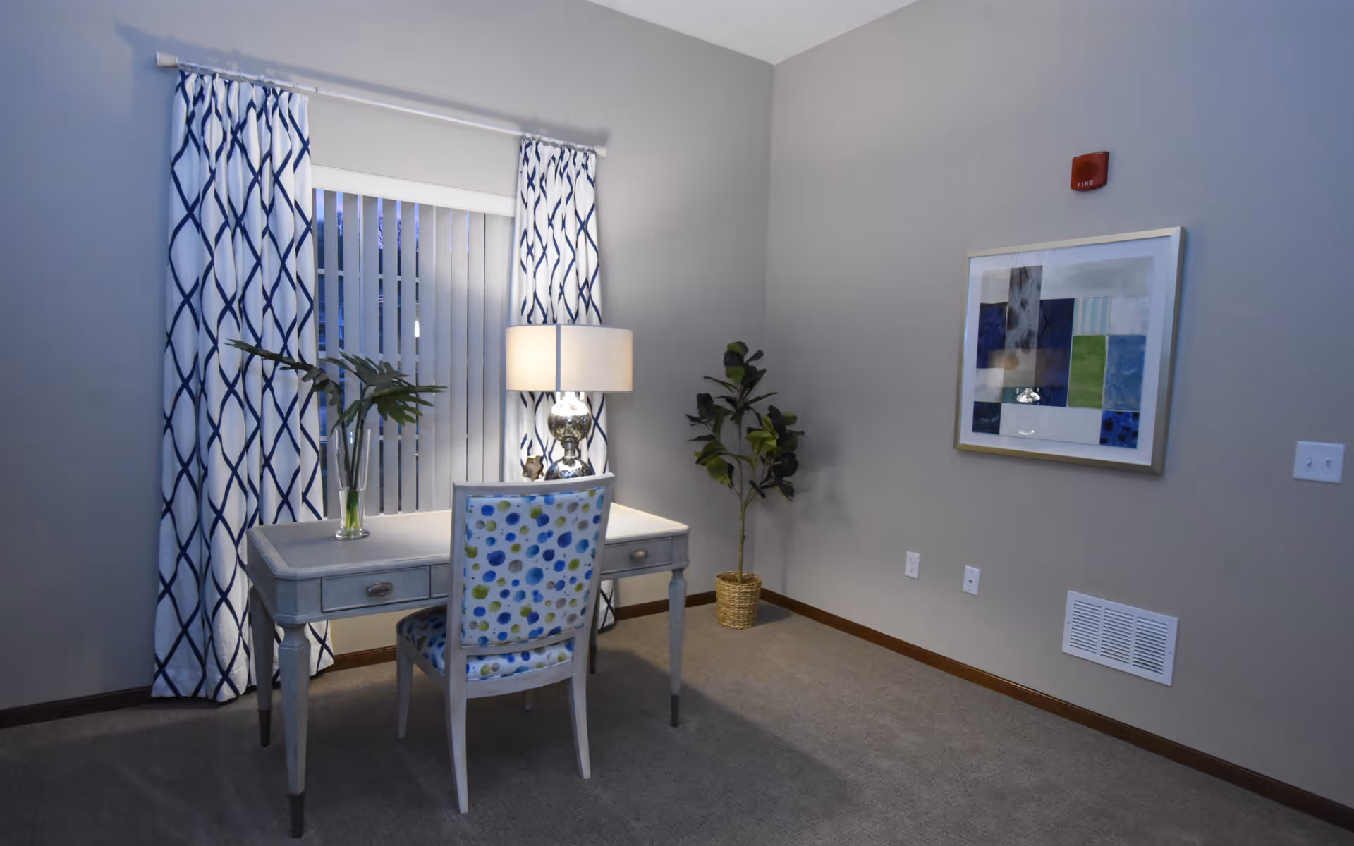 A small, cozy room with a desk and a chair featuring a blue and green polka dot pattern. The desk has a lamp and a glass vase with green leaves. The window behind the desk is covered with vertical blinds and blue and white patterned curtains. A potted plant is placed in the corner, and a framed abstract artwork hangs on the wall. The room has gray walls and carpeted flooring.
