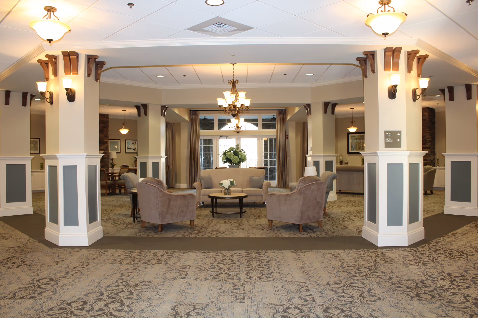 A spacious and well-lit assisted living facility common area with beige walls and carpeted floors featuring a patterned design. The room has multiple columns with decorative wood accents and wall sconces. In the center, there is a seating arrangement consisting of a beige sofa, two matching armchairs, and a round coffee table with a flower vase. Large windows with curtains and multiple chandeliers provide warm lighting.