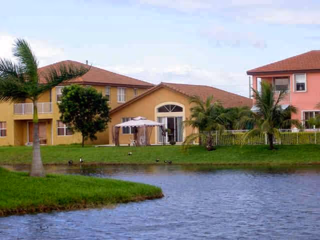 View of a senior living facility with two buildings featuring red-tiled roofs and beige and pink walls, surrounded by palm trees and greenery, with a small lake in the foreground.