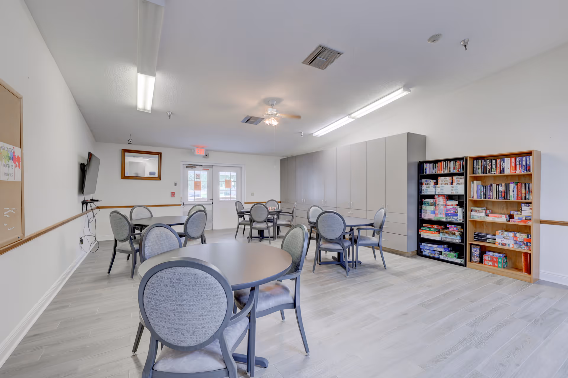 A bright common room with several round tables and chairs arranged for group activities. There are two bookshelves filled with board games and puzzles against the right wall. The room has light-colored flooring, white walls, and a ceiling fan with lights. A wall-mounted TV and a bulletin board are visible on the left wall, and double doors with windows are at the far end of the room.