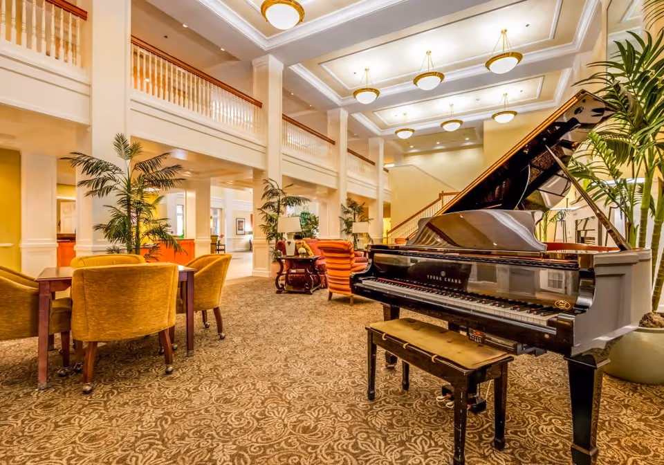 Spacious and elegant common area with a grand piano in the foreground, surrounded by comfortable seating including yellow chairs and orange armchairs. The room features high ceilings with multiple round ceiling lights, large potted plants, and a patterned carpet. A staircase and upper balcony railing are visible in the background.