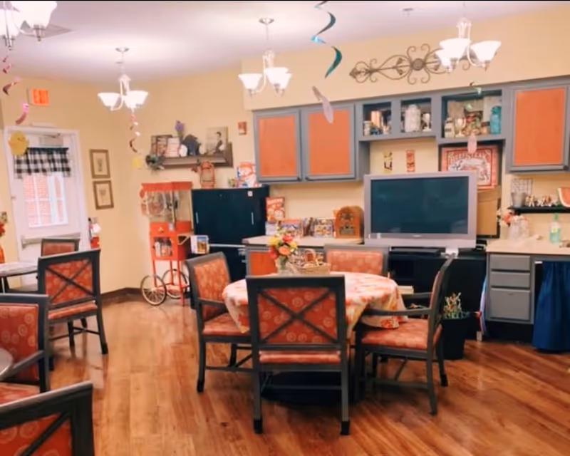 Communal dining room with round tables and upholstered chairs, a TV and kitchenette cabinets against the back wall and hanging decorations.