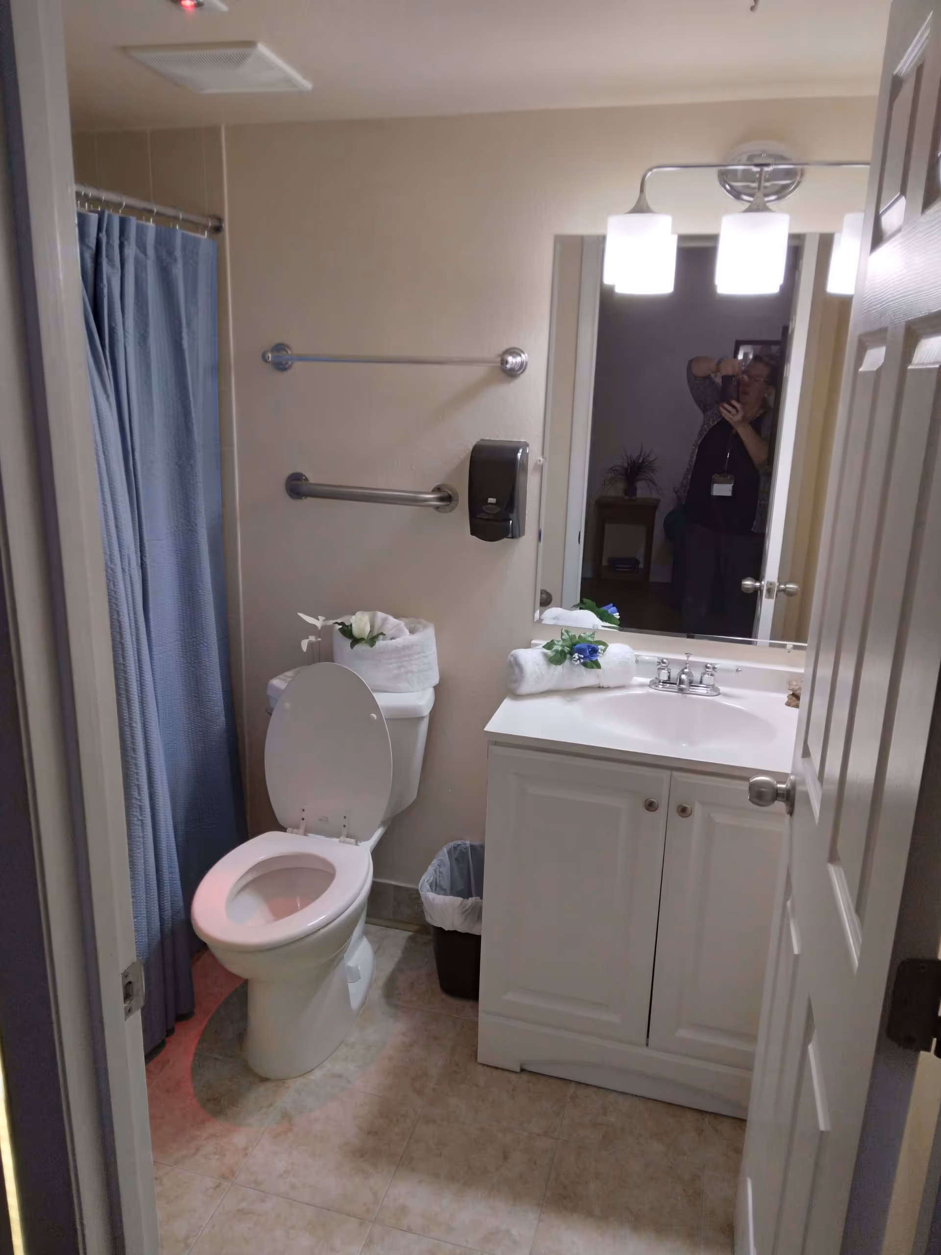 A small bathroom with a white toilet, a white vanity with a sink, and a mirror above it. The toilet lid is open, and there are rolled white towels with decorative flowers on the toilet tank and vanity. A blue shower curtain is visible to the left. The floor is tiled, and there is a trash can next to the vanity. A person is partially visible in the mirror taking the photo.