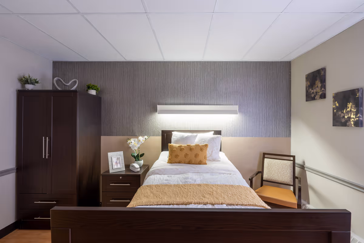 A single-bed bedroom in a senior living facility with dark wood furniture, a nightstand, wardrobe, chair, and decorative pillows.