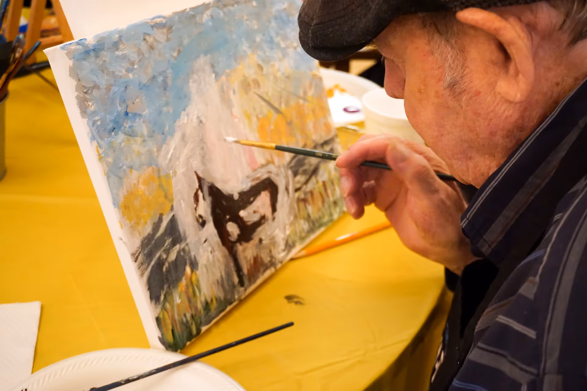An elderly man wearing a dark cap and striped shirt is painting on a canvas. The painting appears to depict a white horse with a black patch on its body, set against a background of blue sky and yellow foliage. The man is holding a paintbrush close to the canvas, which is propped up on a yellow table covered with art supplies.